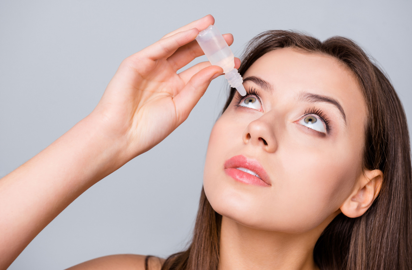 A woman holding a small bottle of eye drops in her right hand and putting them on her right eye.