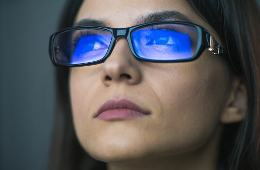 A close-up of a woman with blue light reflected on her glasses from a screen